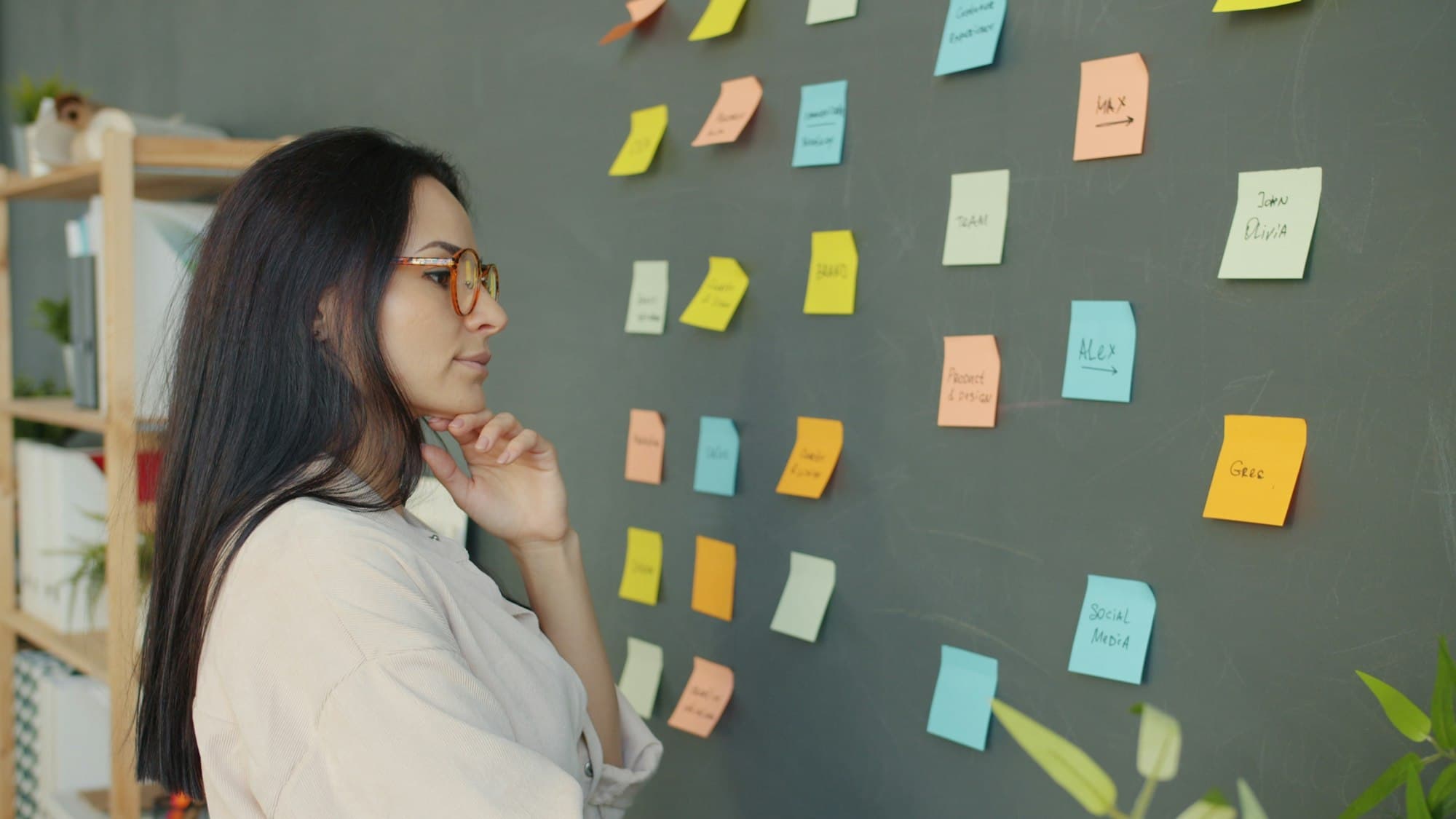 Une femme à lunettes, la main sous le menton, réfléchit devant un mur couvert de post-it de couleur.