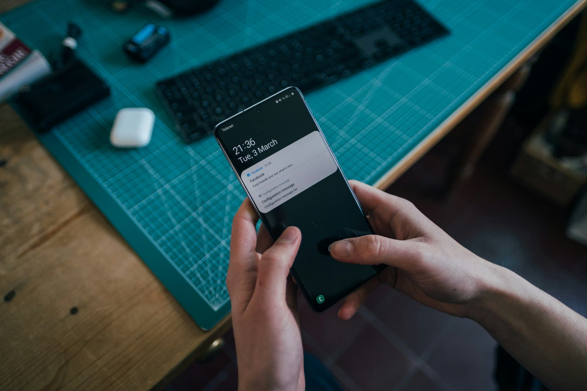 Des mains tiennent un smartphone qui affiche une notification, au-dessus d'un bureau avec un clavier et des écouteurs.