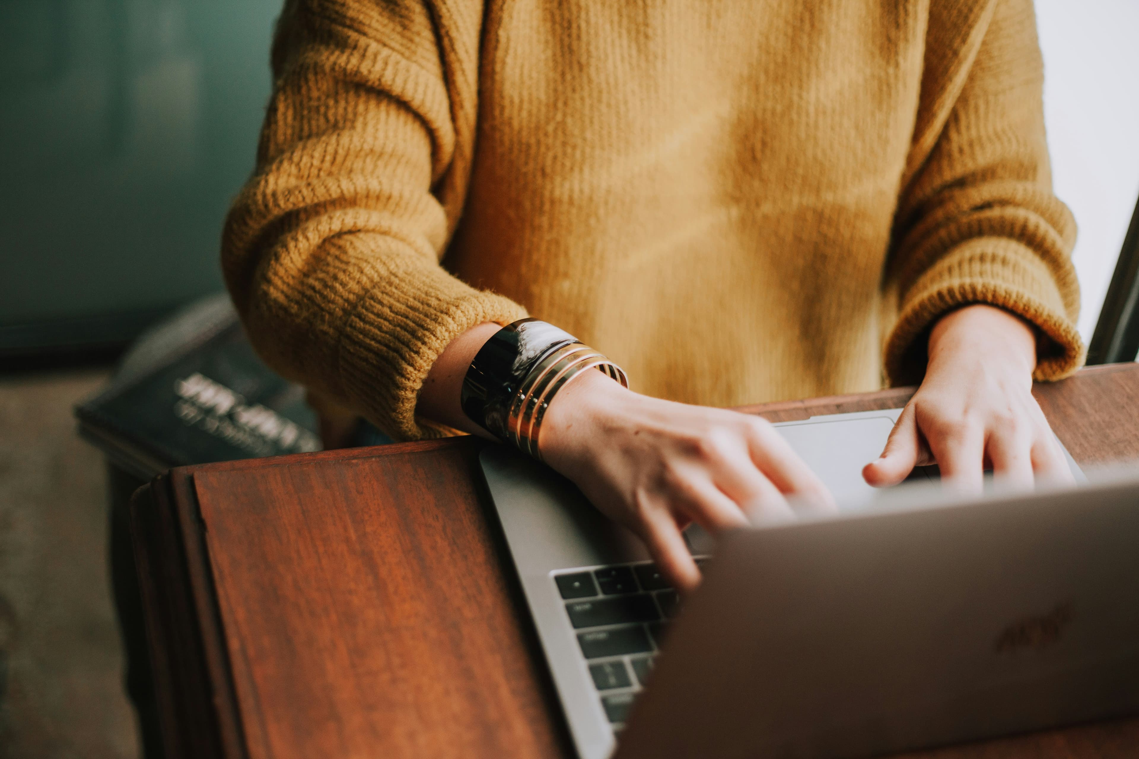 Une personne en pull moutarde tape sur un ordinateur portable posé sur un bureau en bois.